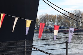 Colorful triangular flags are hanging on a string, with a large dark structure in the background. The setting appears to be near a body of water, with trees and small buildings visible across the water. The flags have various patterns and colors, including yellow, black, white, and red.