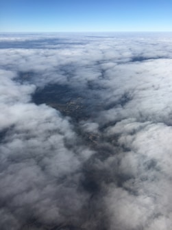 Aerial view showcasing a vast expanse of fluffy clouds, with some open spaces revealing glimpses of the land below. The horizon displays a clear blue sky merging with the thick cloud layer.