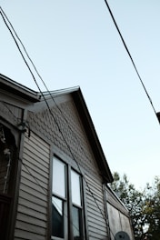 A partial view of a house with a light brown wooden exterior. The house features two windows with visible trim, and the upper part of the siding has a distinct pattern of fish scale shingles. Multiple electrical or utility lines are crossing diagonally in front of the house. The sky is clear and light blue, and some tree foliage is partially visible on the right side of the image.