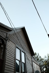 A partial view of a house with a light brown wooden exterior. The house features two windows with visible trim, and the upper part of the siding has a distinct pattern of fish scale shingles. Multiple electrical or utility lines are crossing diagonally in front of the house. The sky is clear and light blue, and some tree foliage is partially visible on the right side of the image.