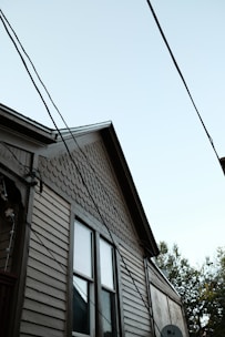 A partial view of a house with a light brown wooden exterior. The house features two windows with visible trim, and the upper part of the siding has a distinct pattern of fish scale shingles. Multiple electrical or utility lines are crossing diagonally in front of the house. The sky is clear and light blue, and some tree foliage is partially visible on the right side of the image.