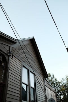 A partial view of a house with a light brown wooden exterior. The house features two windows with visible trim, and the upper part of the siding has a distinct pattern of fish scale shingles. Multiple electrical or utility lines are crossing diagonally in front of the house. The sky is clear and light blue, and some tree foliage is partially visible on the right side of the image.