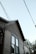 A partial view of a house with a light brown wooden exterior. The house features two windows with visible trim, and the upper part of the siding has a distinct pattern of fish scale shingles. Multiple electrical or utility lines are crossing diagonally in front of the house. The sky is clear and light blue, and some tree foliage is partially visible on the right side of the image.