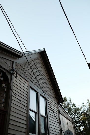 A partial view of a house with a light brown wooden exterior. The house features two windows with visible trim, and the upper part of the siding has a distinct pattern of fish scale shingles. Multiple electrical or utility lines are crossing diagonally in front of the house. The sky is clear and light blue, and some tree foliage is partially visible on the right side of the image.