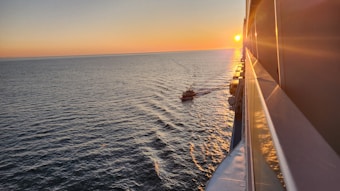 The image captures a serene ocean scene at sunset with a small boat navigating through the water. The sun is low on the horizon, casting a warm golden glow across the sea's surface. The edge of a large vessel is visible on the right side of the image, reflecting the sunlight.