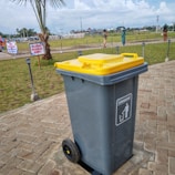 A large gray garbage bin with a yellow lid is placed on a paved pathway in a grassy outdoor area. The bin has wheels at the base and a label indicating its use for trash. In the background, there are people walking, a palm tree, signs with text in another language, and a cloudy sky.