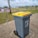 A large gray garbage bin with a yellow lid is placed on a paved pathway in a grassy outdoor area. The bin has wheels at the base and a label indicating its use for trash. In the background, there are people walking, a palm tree, signs with text in another language, and a cloudy sky.