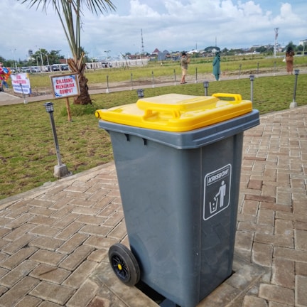 A large gray garbage bin with a yellow lid is placed on a paved pathway in a grassy outdoor area. The bin has wheels at the base and a label indicating its use for trash. In the background, there are people walking, a palm tree, signs with text in another language, and a cloudy sky.