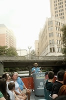 A group of people sitting on a river tour boat, with a guide standing at the front wearing a hat. They are on a city river with tall buildings and a bridge in the background.
