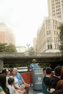 A group of people sitting on a river tour boat, with a guide standing at the front wearing a hat. They are on a city river with tall buildings and a bridge in the background.