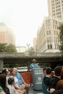 A group of people sitting on a river tour boat, with a guide standing at the front wearing a hat. They are on a city river with tall buildings and a bridge in the background.
