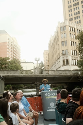 A group of people sitting on a river tour boat, with a guide standing at the front wearing a hat. They are on a city river with tall buildings and a bridge in the background.