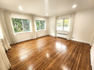 A spacious, unfurnished room with polished hardwood floors and bright white walls. Large windows with sheer curtains allow natural light to illuminate the space. Outside, green foliage is visible through the windows, adding a touch of nature to the scene.