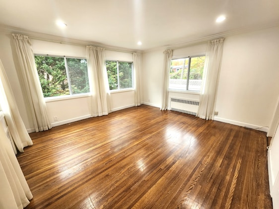 A spacious, unfurnished room with polished hardwood floors and bright white walls. Large windows with sheer curtains allow natural light to illuminate the space. Outside, green foliage is visible through the windows, adding a touch of nature to the scene.