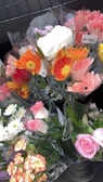 Several bouquets of colorful flowers are wrapped in transparent plastic, displayed in a metal container. The bouquets include a variety of flowers such as pink roses, orange gerberas, white chrysanthemums, and yellow daisies, creating a vibrant assortment.