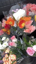Several bouquets of colorful flowers are wrapped in transparent plastic, displayed in a metal container. The bouquets include a variety of flowers such as pink roses, orange gerberas, white chrysanthemums, and yellow daisies, creating a vibrant assortment.