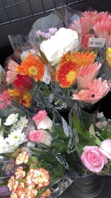 Several bouquets of colorful flowers are wrapped in transparent plastic, displayed in a metal container. The bouquets include a variety of flowers such as pink roses, orange gerberas, white chrysanthemums, and yellow daisies, creating a vibrant assortment.
