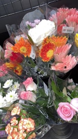 Several bouquets of colorful flowers are wrapped in transparent plastic, displayed in a metal container. The bouquets include a variety of flowers such as pink roses, orange gerberas, white chrysanthemums, and yellow daisies, creating a vibrant assortment.