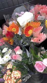 Several bouquets of colorful flowers are wrapped in transparent plastic, displayed in a metal container. The bouquets include a variety of flowers such as pink roses, orange gerberas, white chrysanthemums, and yellow daisies, creating a vibrant assortment.