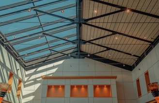A modern architectural interior featuring a large glass ceiling with metal beams, allowing natural light to illuminate the space below. The walls are adorned with square panels in neutral tones, and there are recessed lighting fixtures. A decorative element resembling a basket-like structure is present.