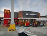 A McDonald's restaurant with its distinct logo is visible, featuring a modern design with a combination of red, yellow, and brown elements. The entrance is decorated with flower arrangements and a large red heart. Several motorcycles and a tricycle are parked in front, and a tent is set up nearby.