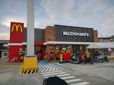 A McDonald's restaurant with its distinct logo is visible, featuring a modern design with a combination of red, yellow, and brown elements. The entrance is decorated with flower arrangements and a large red heart. Several motorcycles and a tricycle are parked in front, and a tent is set up nearby.
