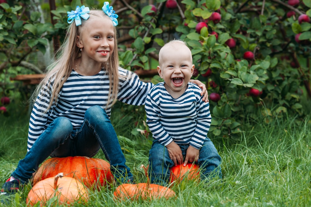 a boy and girl sitting on pumpkins in a grassy area
