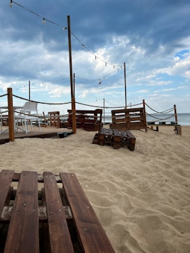 A sandy beach area with wooden pallets and simple outdoor furniture arranged for seating. String lights hang above, adding a cozy and ambient atmosphere. The sky is partly cloudy, suggesting a relaxed coastal setting.