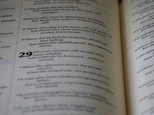 A close-up view of an open book displaying text in two columns. The text appears to be religious or philosophical in nature, with verses numbered and formatted in a poetic structure.