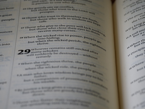 A close-up view of an open book displaying text in two columns. The text appears to be religious or philosophical in nature, with verses numbered and formatted in a poetic structure.