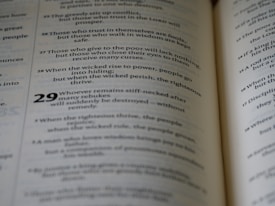A close-up view of an open book displaying text in two columns. The text appears to be religious or philosophical in nature, with verses numbered and formatted in a poetic structure.
