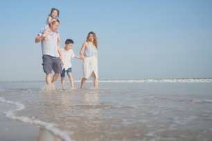 A family of four walks along the beach, enjoying the shallow water. The father carries a young girl on his shoulders, while holding hands with a boy who is walking alongside the mother. The sun shines brightly in the clear blue sky, and gentle waves can be seen in the background.