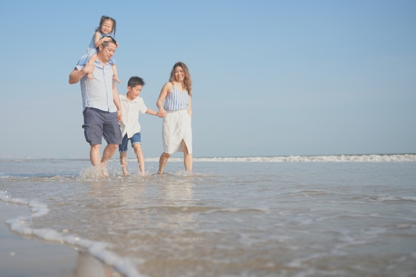 A family of four walks along the beach, enjoying the shallow water. The father carries a young girl on his shoulders, while holding hands with a boy who is walking alongside the mother. The sun shines brightly in the clear blue sky, and gentle waves can be seen in the background.