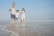 A family of four walks along the beach, enjoying the shallow water. The father carries a young girl on his shoulders, while holding hands with a boy who is walking alongside the mother. The sun shines brightly in the clear blue sky, and gentle waves can be seen in the background.