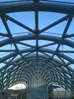 A modern architectural structure featuring a complex, arched design with crisscrossing metal beams against a clear blue sky. The framework has a symmetrical, tunnel-like appearance with visible tension cables, suggesting a bridge or walkway. Below, parts of urban buildings and rooftops are visible at the bottom edge.