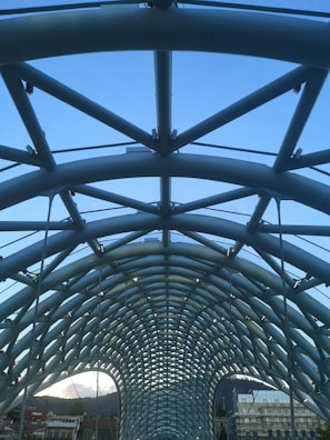A modern architectural structure featuring a complex, arched design with crisscrossing metal beams against a clear blue sky. The framework has a symmetrical, tunnel-like appearance with visible tension cables, suggesting a bridge or walkway. Below, parts of urban buildings and rooftops are visible at the bottom edge.