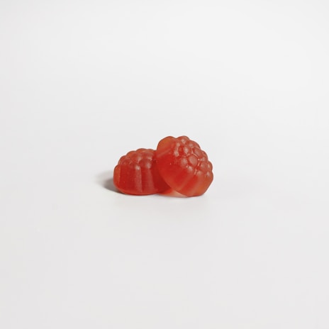 Two red gummy candies shaped like raspberries are placed on a clean, white surface. The candies have a translucent appearance with a glossy finish, displaying a bumpy texture resembling fruit details.