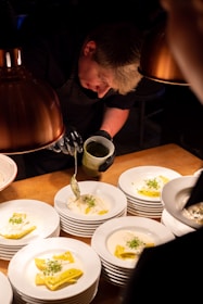 A chef is carefully plating dishes under warm lighting. Each white plate is topped with ravioli in a creamy sauce and garnished with fresh herbs. The chef uses a spoon to add sauce with precision, demonstrating attention to detail and culinary skill.