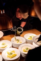 A chef is carefully plating dishes under warm lighting. Each white plate is topped with ravioli in a creamy sauce and garnished with fresh herbs. The chef uses a spoon to add sauce with precision, demonstrating attention to detail and culinary skill.