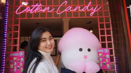 A person smiles while holding a large, pink cotton candy shaped like a character with a face in front of a colorful shop. The shop has a neon sign that reads 'Cotton Candy' and bright posters on the walls.