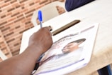 A hand is writing with a blue pen on a piece of paper that appears to be a magazine or newspaper. The publication features a photograph of a smiling person. A smartphone and a brick-patterned wall are visible in the background.