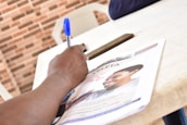 A hand is writing with a blue pen on a piece of paper that appears to be a magazine or newspaper. The publication features a photograph of a smiling person. A smartphone and a brick-patterned wall are visible in the background.