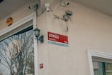 A beige wall displays a red street sign with the words 'Dönüş Sokak' and directional information. The wall has various electrical components and security cameras attached. A classic-style outdoor lantern is mounted nearby. Reflecting on the glass of the adjacent building is a large tree without leaves.