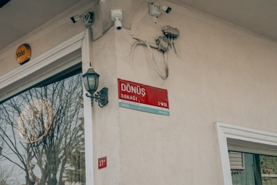 A beige wall displays a red street sign with the words 'Dönüş Sokak' and directional information. The wall has various electrical components and security cameras attached. A classic-style outdoor lantern is mounted nearby. Reflecting on the glass of the adjacent building is a large tree without leaves.