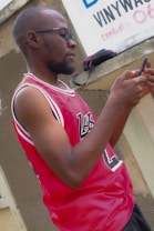 A person is standing outdoors, wearing sunglasses and a red sports jersey with white and black lettering. They are holding and looking at a smartphone. In the background, there's a concrete wall with a partially visible sign and a black-and-pink bag resting on a ledge.