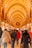 A bustling indoor market scene featuring a curved, ornately decorated ceiling with arches and intricate patterns. The passage is lined with various shops displaying goods, and numerous people are walking along, some carrying bags. Turkish flags are prominently displayed throughout the market.