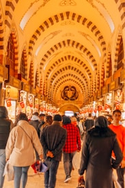 A bustling indoor market scene featuring a curved, ornately decorated ceiling with arches and intricate patterns. The passage is lined with various shops displaying goods, and numerous people are walking along, some carrying bags. Turkish flags are prominently displayed throughout the market.