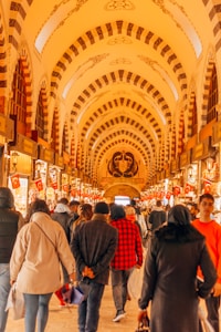 A bustling indoor market scene featuring a curved, ornately decorated ceiling with arches and intricate patterns. The passage is lined with various shops displaying goods, and numerous people are walking along, some carrying bags. Turkish flags are prominently displayed throughout the market.