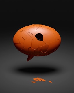 A broken orange speech bubble appears to be floating in mid-air against a dark background. The bubble's surface is cracked, with a section opening into a hollow interior, and a few fragments lie on the ground beneath it.