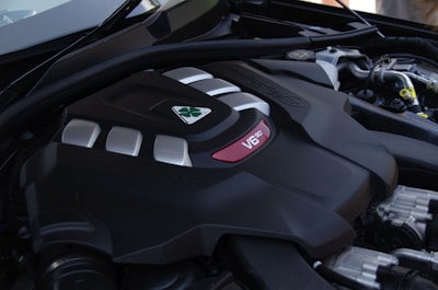 A close-up view of a car engine, prominently featuring a V6 configuration. The engine has a sleek black casing with shiny metallic accents and a distinct triangular emblem. Various components and pipes of the engine system can be seen in the background.